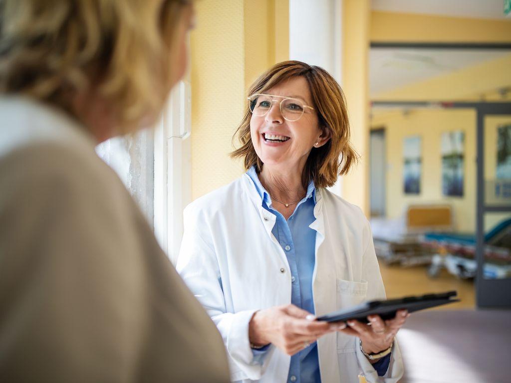 Bild: Ärztin mit Brille und Klemmbrett lächelt ihre Patientin an.