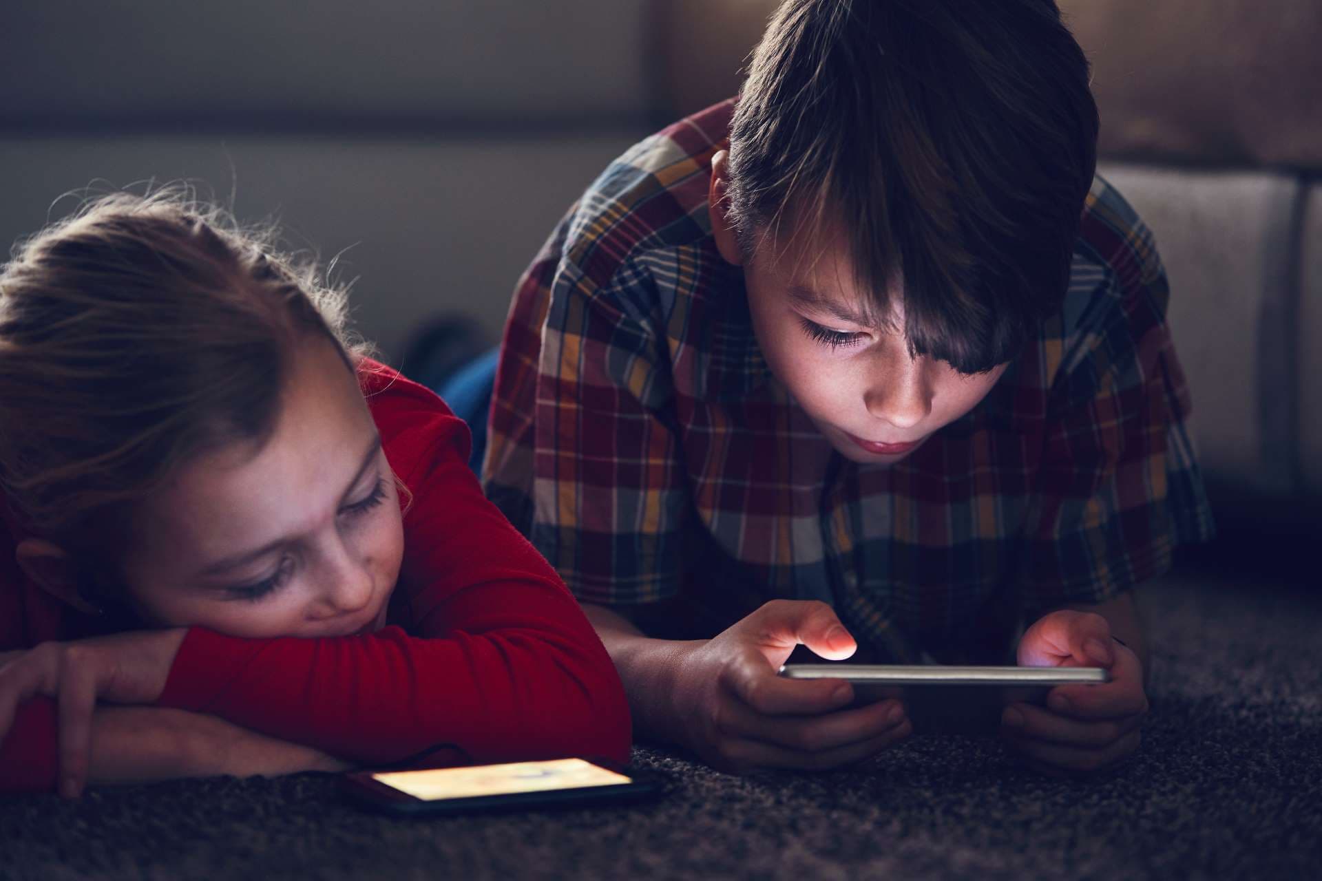 Mediensucht: Ein Mädchen und ein Junge liegen in einem dunklen Zimmer auf dem Boden und schauen müde auf ihr Smartphone.