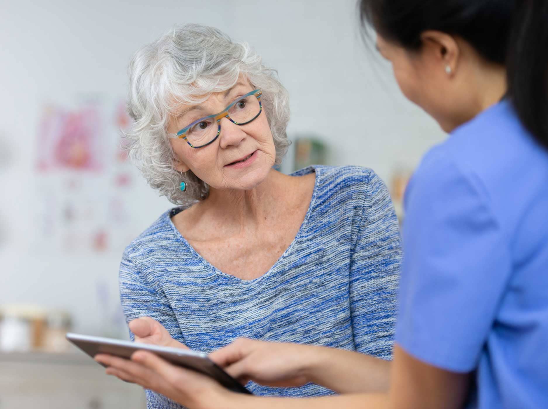 Eine ältere Frau spricht mit ihrer Ärztin und schaut sie dabei an. Die Ärztin hält ein Tablet in der Hand.