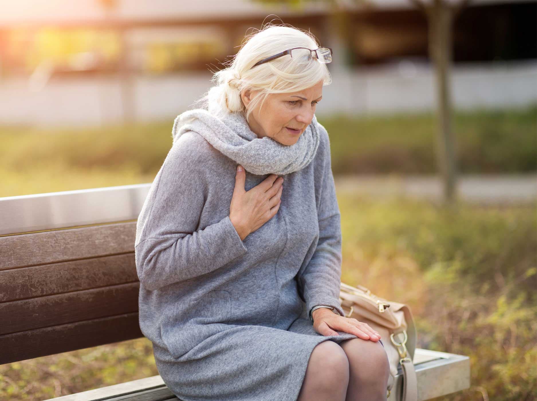 Ältere Frau sitzt draußen auf einer Bank und fasst sich mit einer Hand an die Brust.