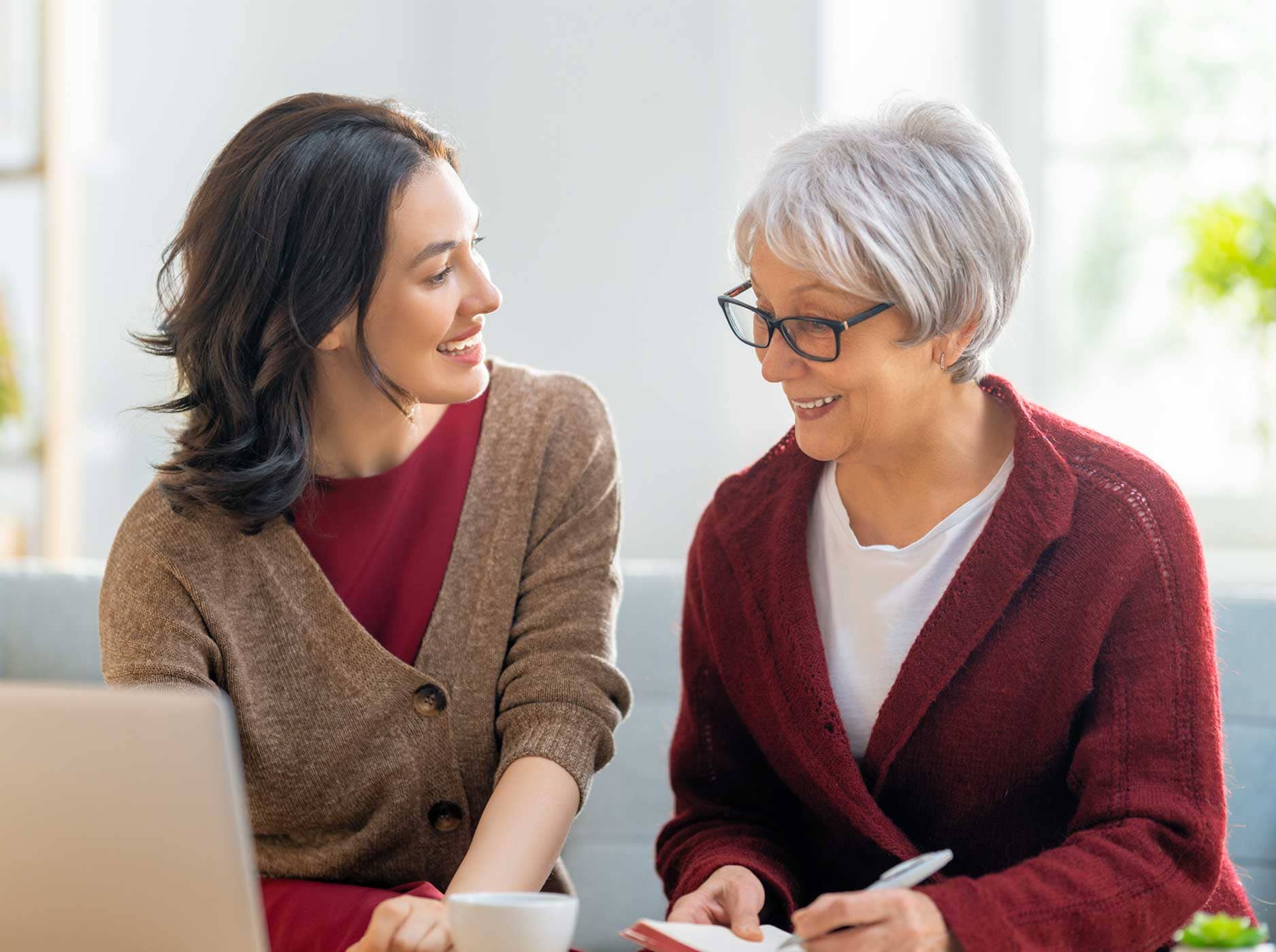 Zwei Frauen sitzen vor einem Notebook und besprechen etwas. Die ältere Frau macht sich Notizen.