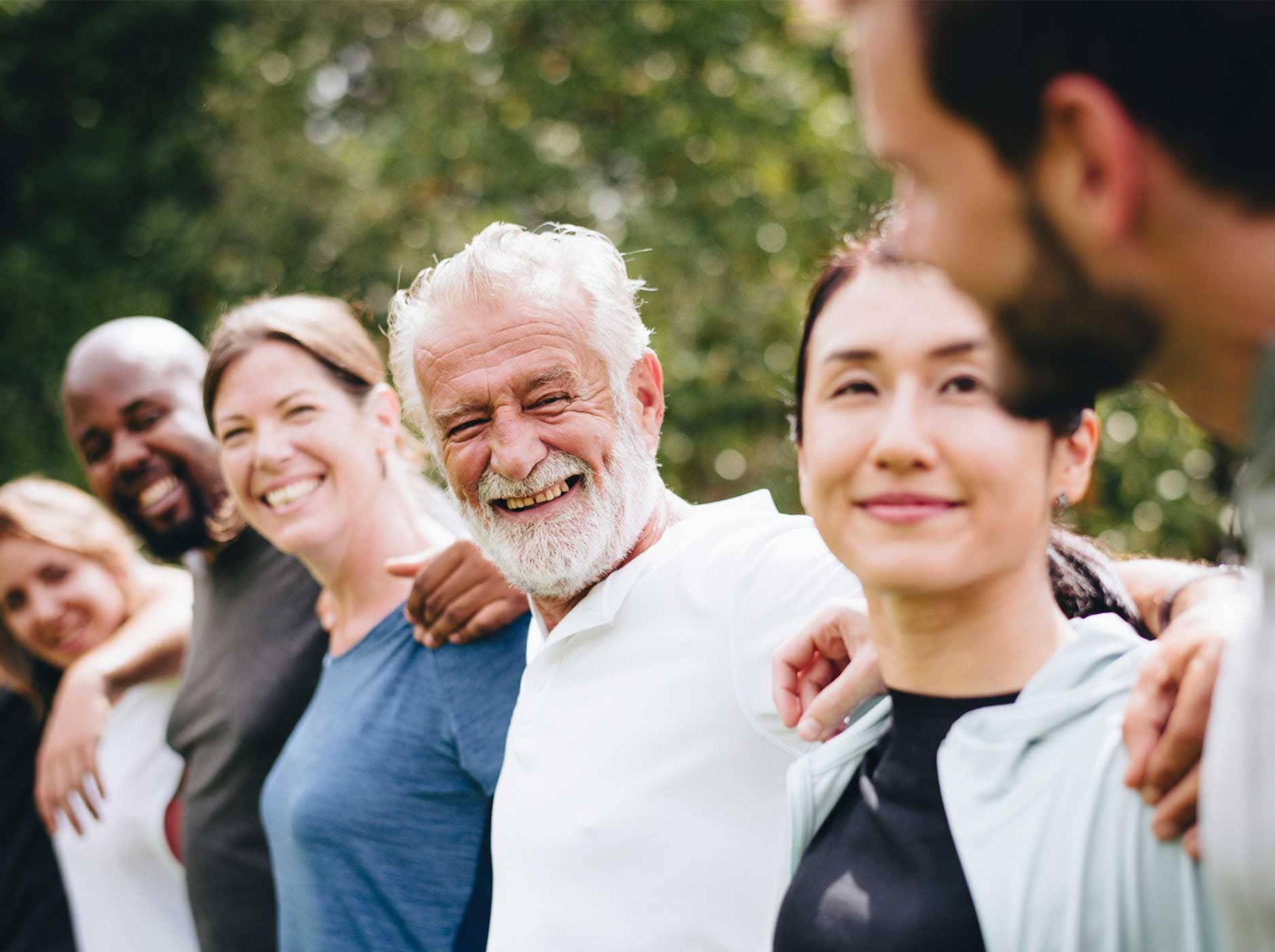 Mehrere lachende Menschen unterschiedlichen Alters stehen in einem Park nebeneinander und bilden eine Menschenkette.