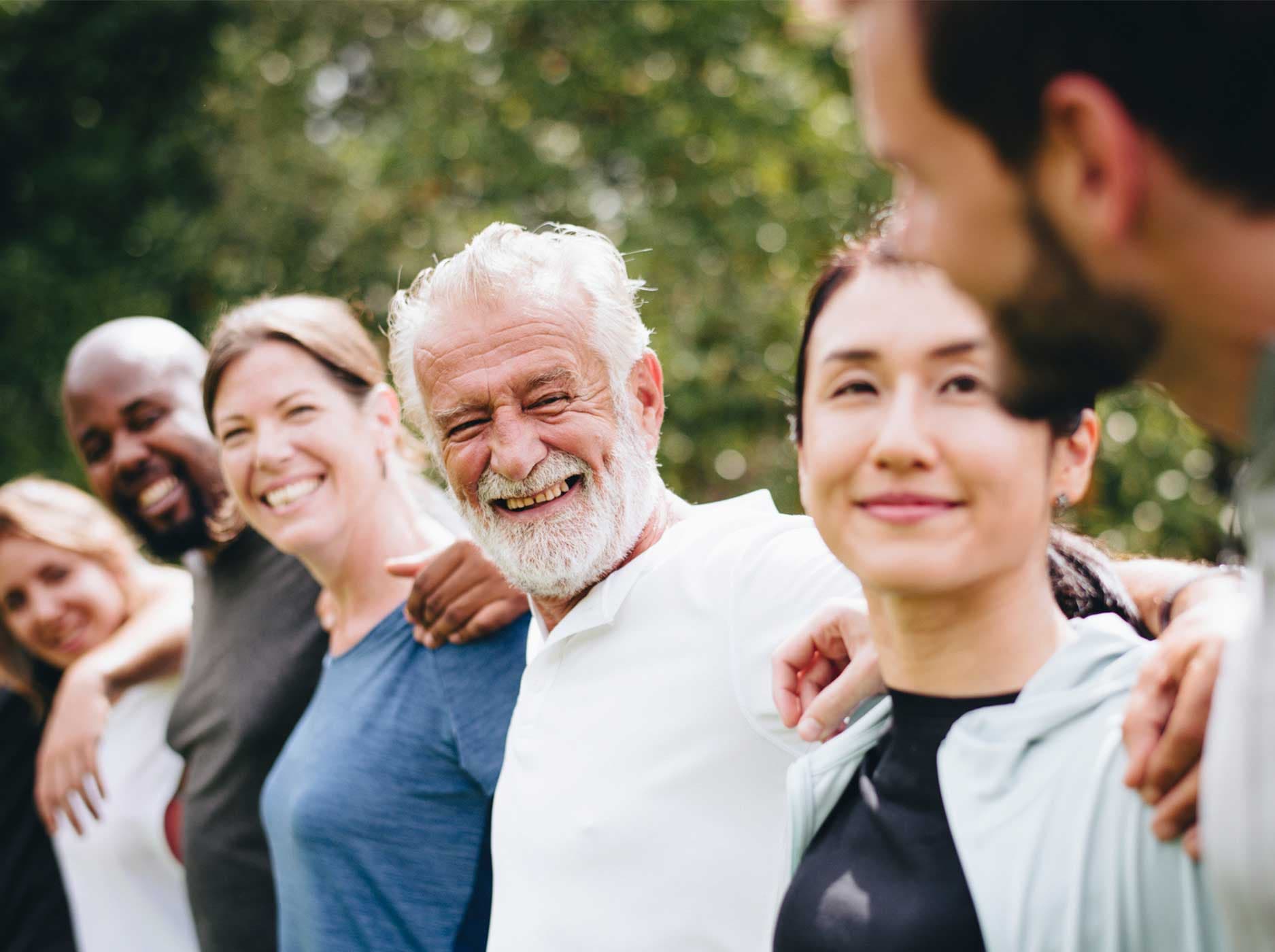 Mehrere lachende Menschen unterschiedlichen Alters stehen in einem Park nebeneinander und bilden eine Menschenkette.