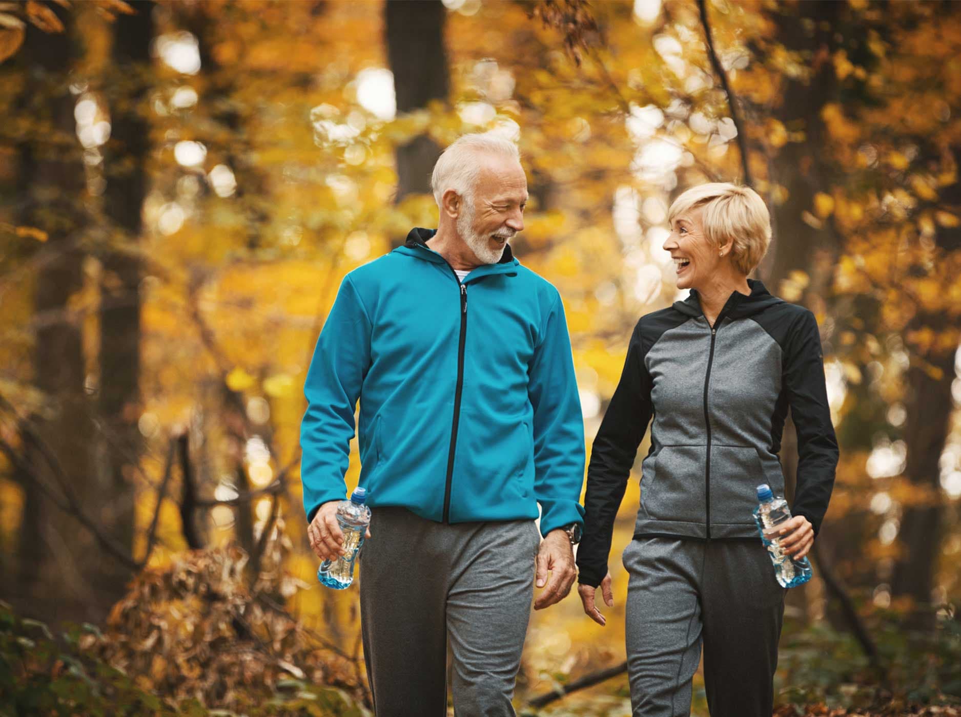 Ein älterer Mann und eine ältere Frau gehen vergnügt in einem herbstlichen Wald in Sportkleidung spazieren.