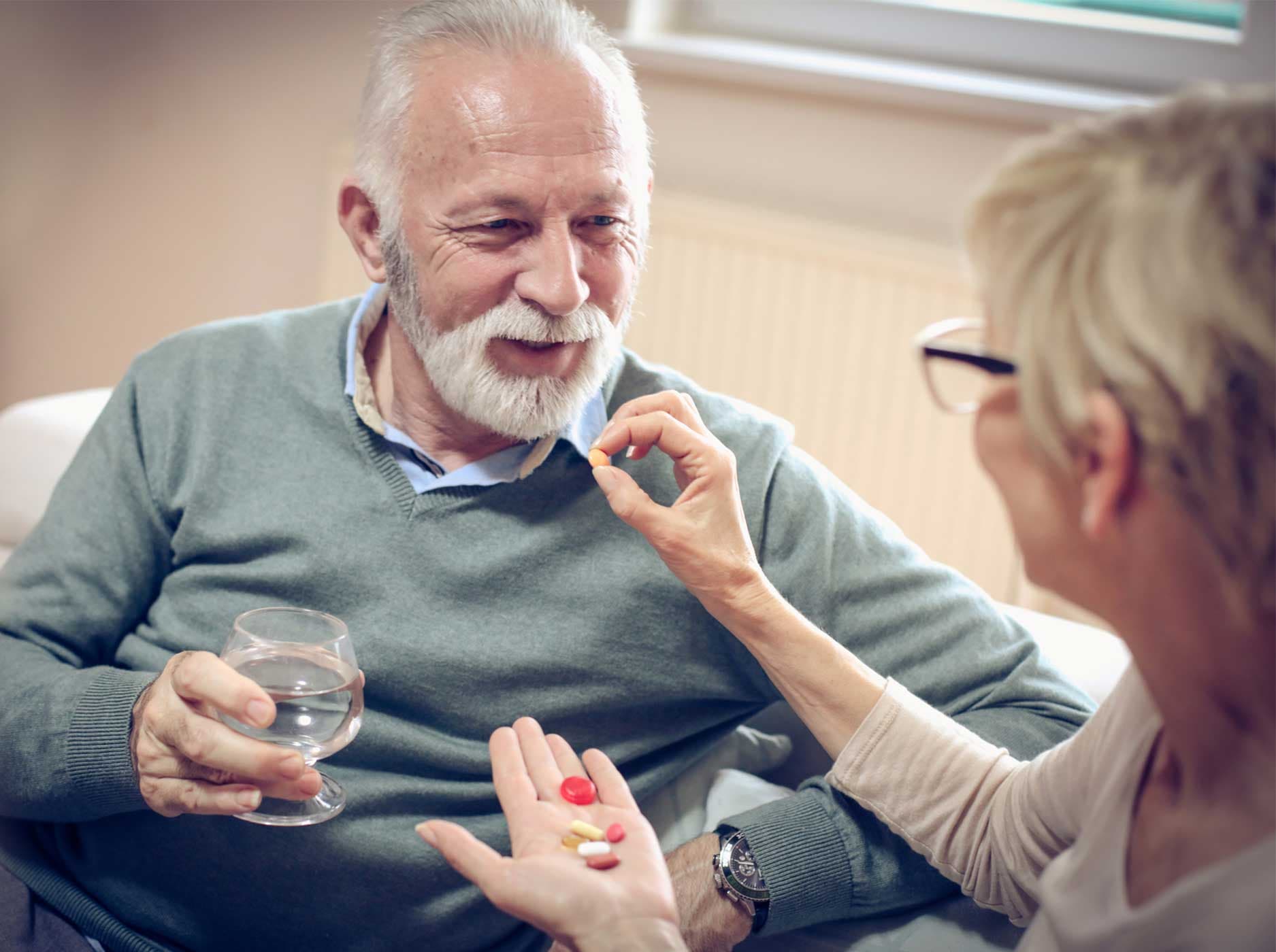 Älteres Paar sitzt entspannt auf einem Sofa. Sie reicht ihm seine Tabletten an.