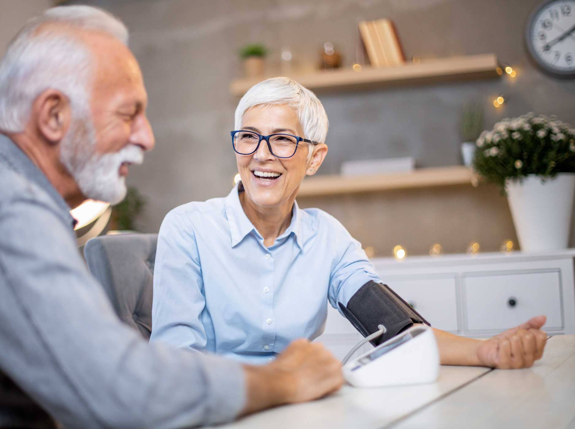 Älterer Mann misst bei seiner Frau zuhause am Esstisch den Blutdruck.