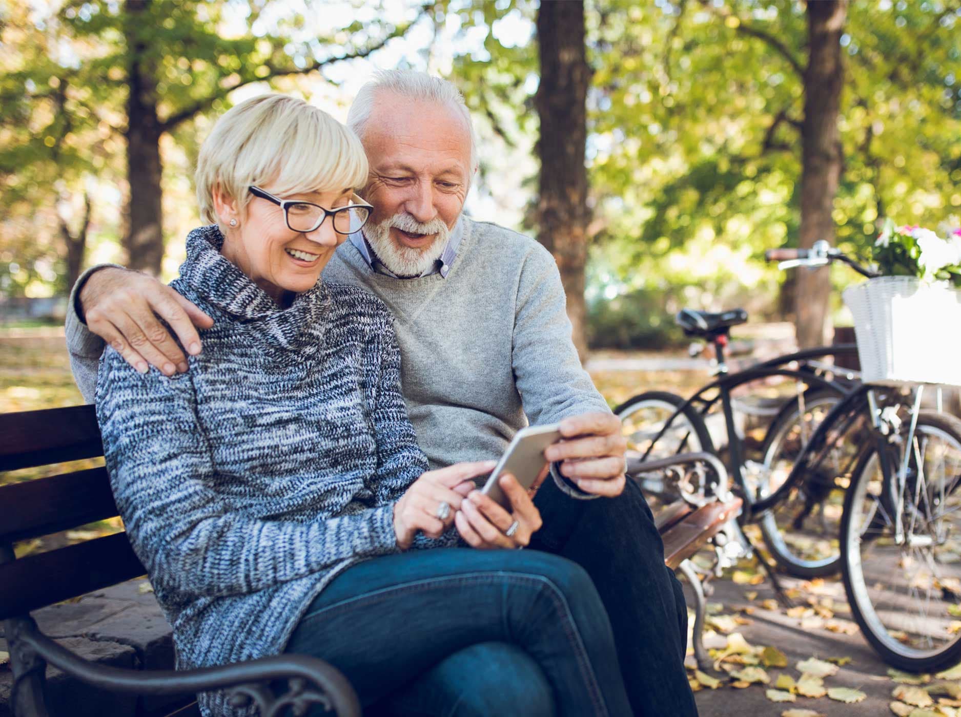 Älteres Paar sitzt vergnügt auf einer Parkbank und schaut gemeinsam auf ein Smartphone. Im Hintergrund stehen zwei Fahrräder.