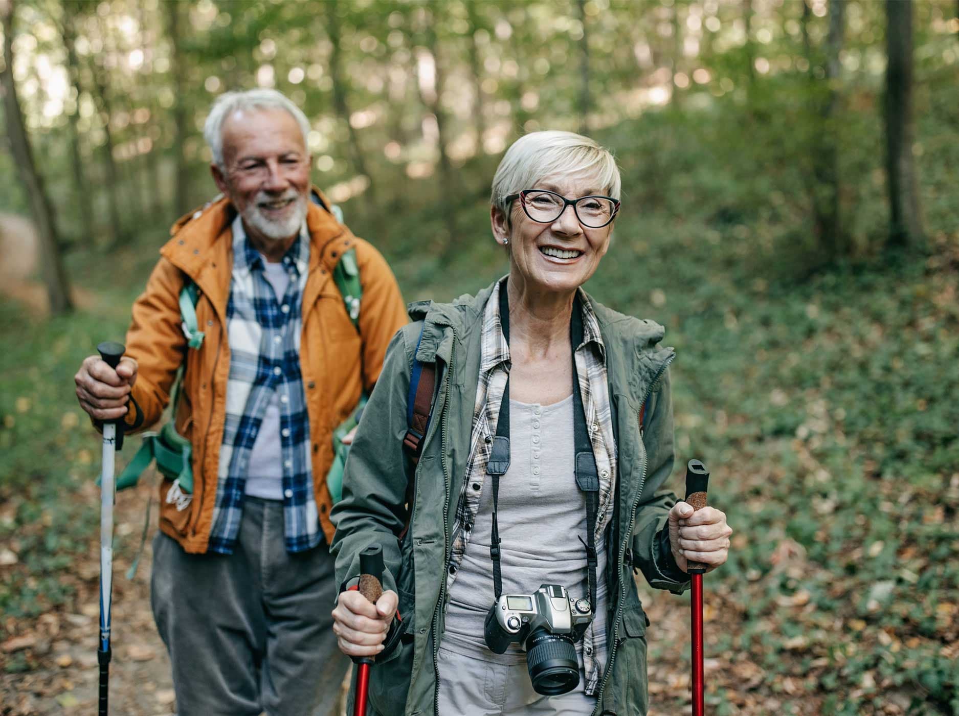 Älteres Paar läuft mit Nordic-Walking-Stöcken durch einen Wald.