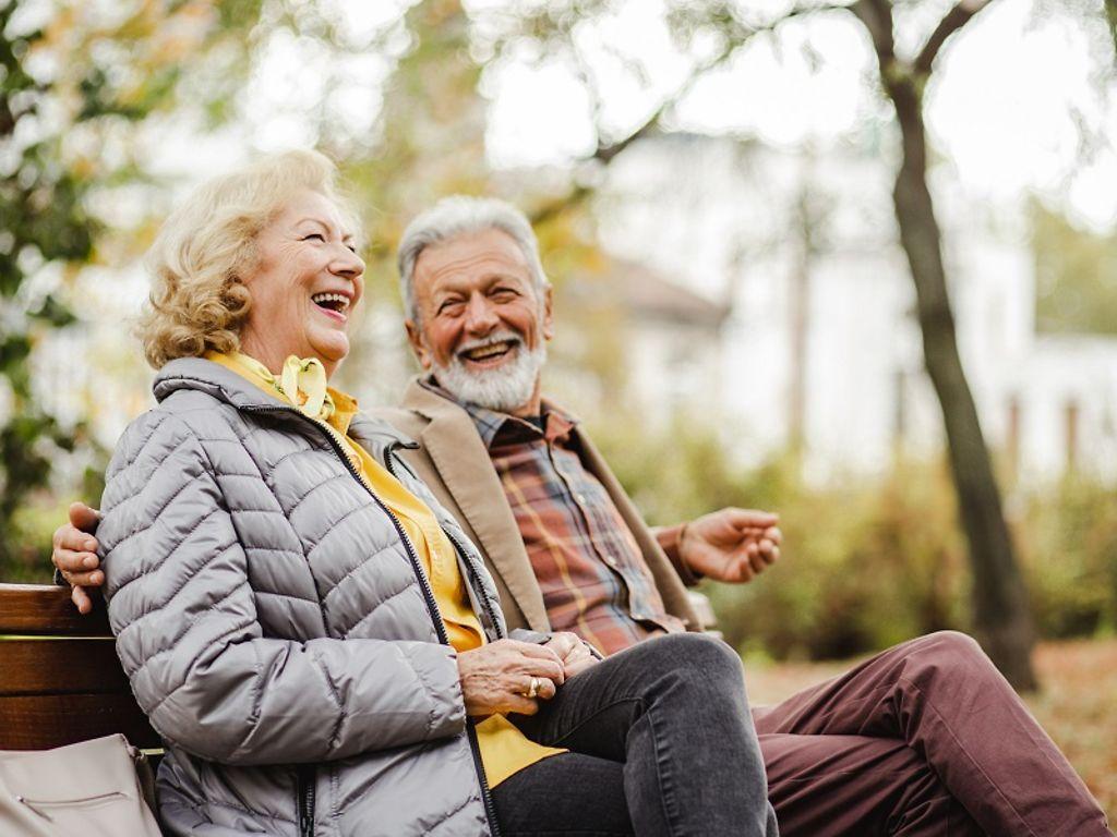 Krankenversicherung für Senioren: Älteres Ehepaar sitzt lachend auf Parkbank