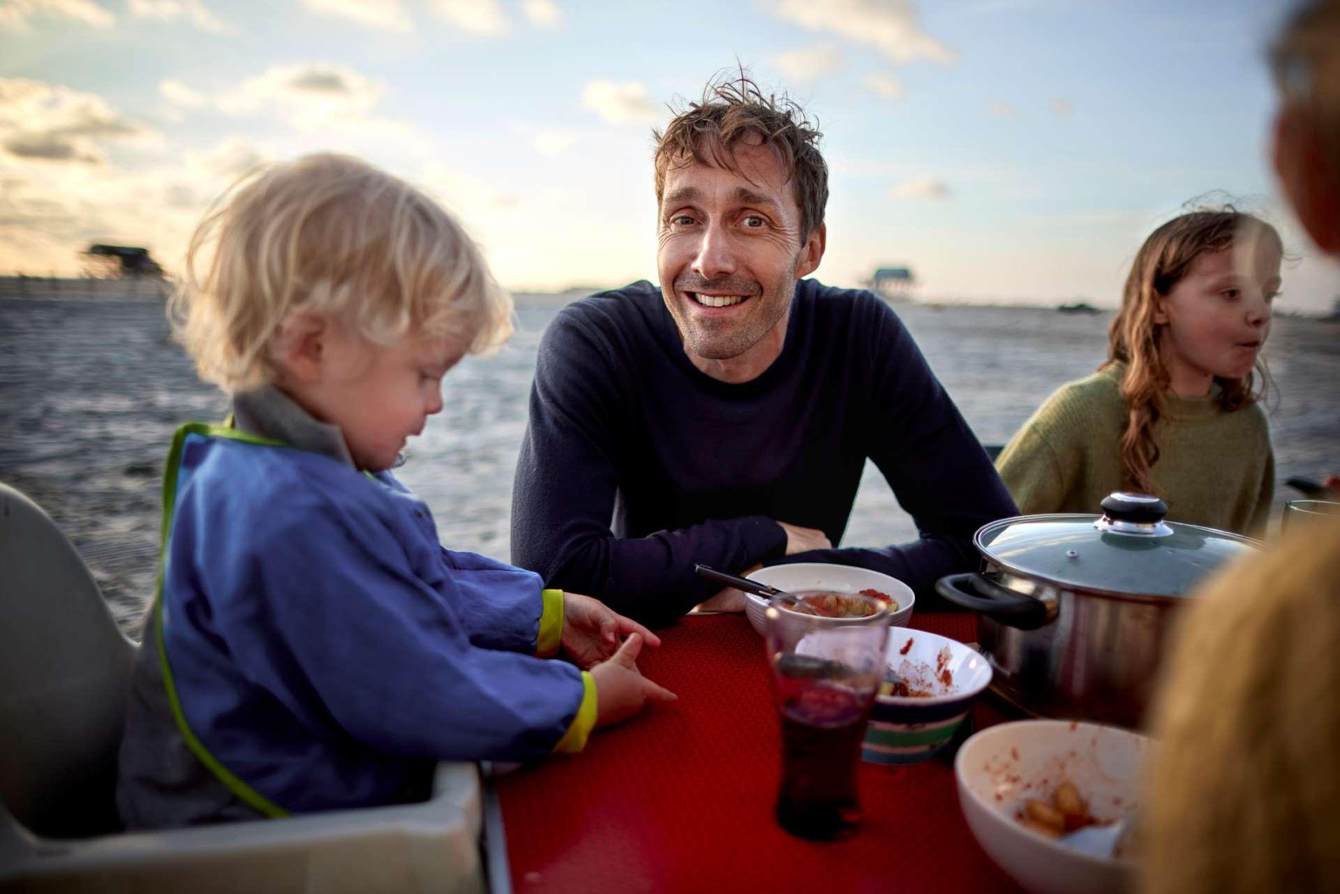 Junge Familie sitzt am Meer an einem Camping-Tisch und isst.
