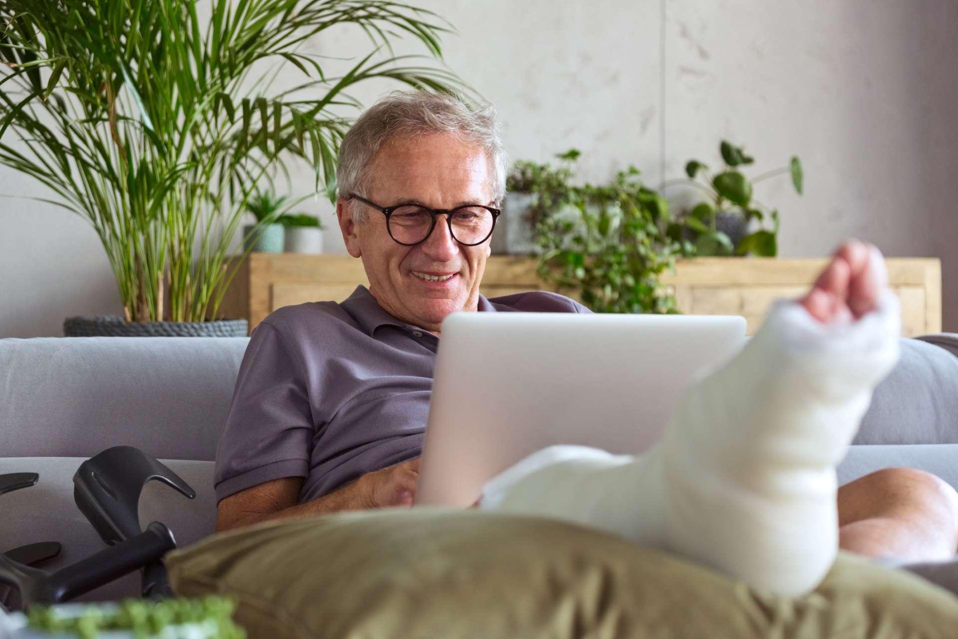 DAK-Hilfsmittellotse: Älterer Mann mit Gipsbein sitz auf dem Sofa und schaut auf sein Notebook.