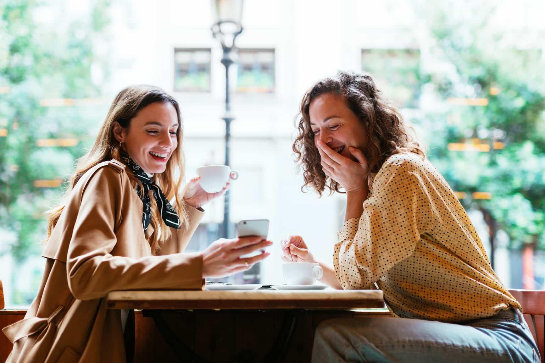 Fragen Bonusprogramm: Zwei Frauen in einem Café, schauen auf ein Handy und haben Spaß.