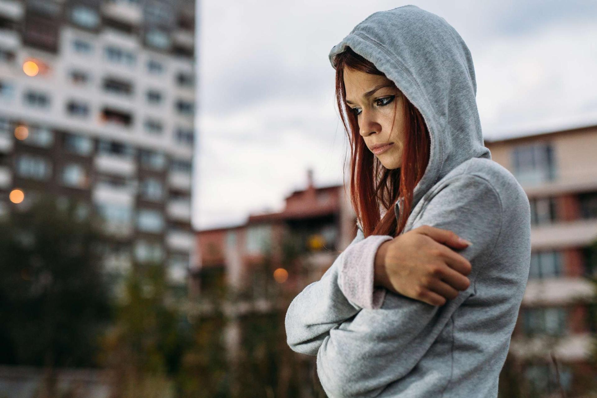 Junges Mädchen mit Kapuzen-Pullover steht draußen in einer Hochhaussiedlung und schaut deprimiert auf den Boden.