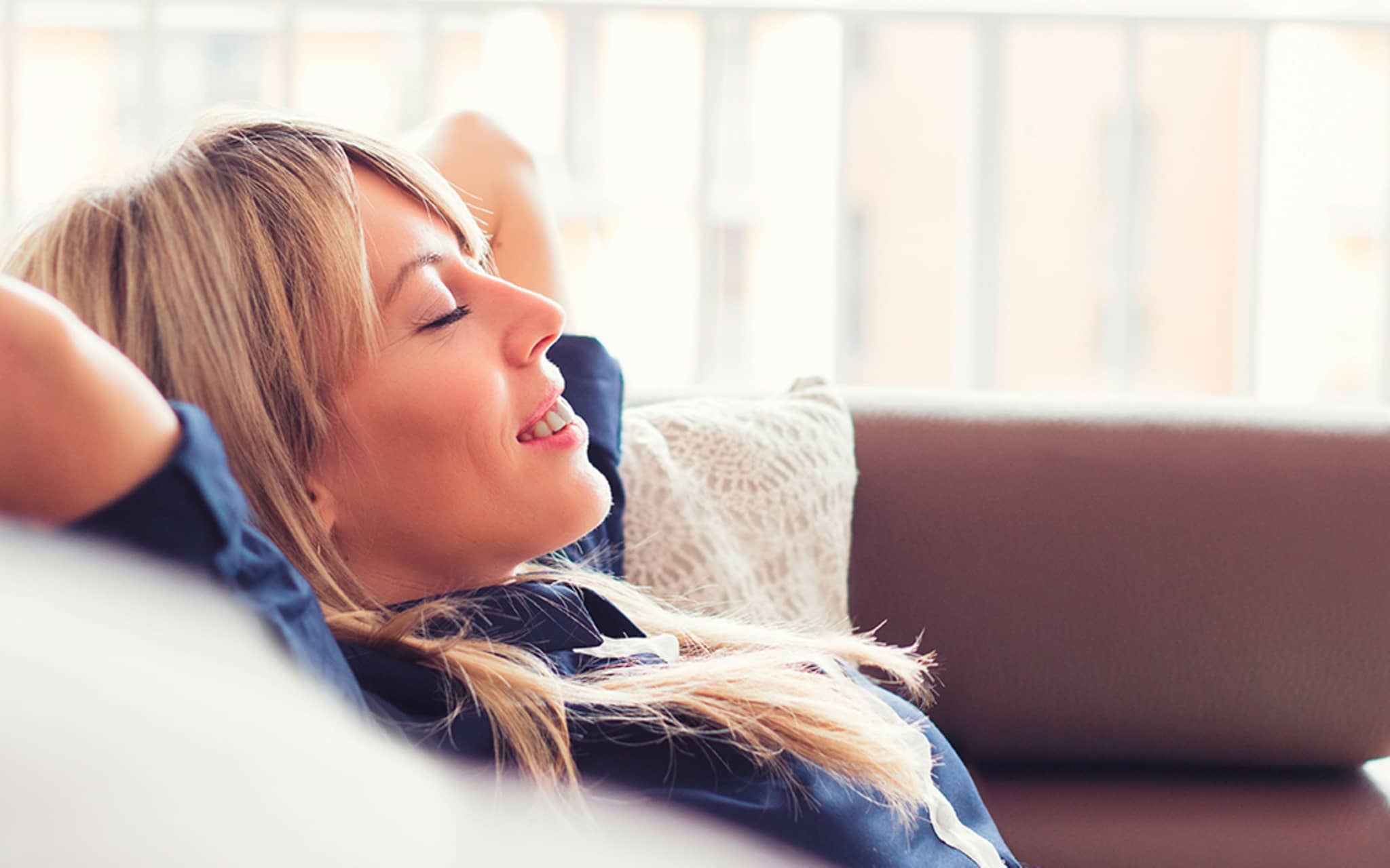Junge Frau verschränkt entspannt die Arme hinter dem Kopf und lehnt sich auf Couch zurück