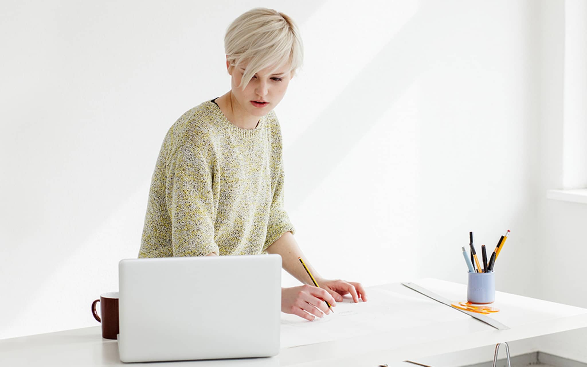 Junge Frau zeichnet konzentriert mit einem Bleistift und schaut auf ihre Vorlage auf dem Laptop