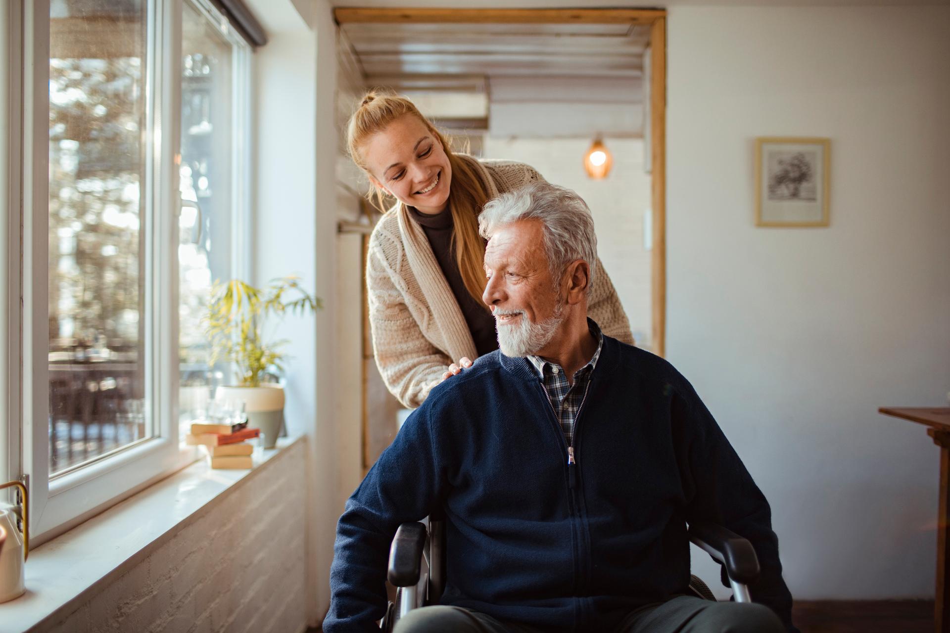 Pflegeleistungen: Junge Frau steht hinter älteren Mann im Rollstuhl.