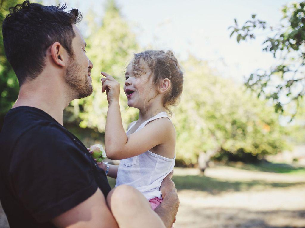 Krankenversicherung für Familien: Junger Vater hält draußen verspielt seine Tochter im Arm