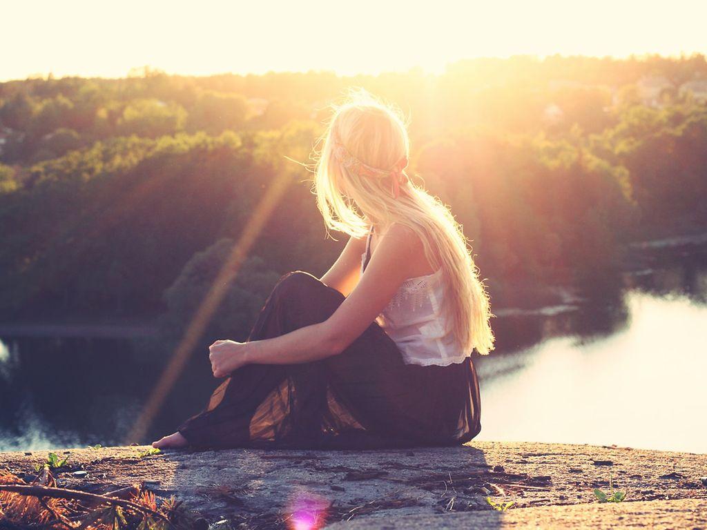 Bild: PlusGesundheit: Blonde Frau sitzt auf einem Felsen und schaut dem Sonnenuntergang zu.