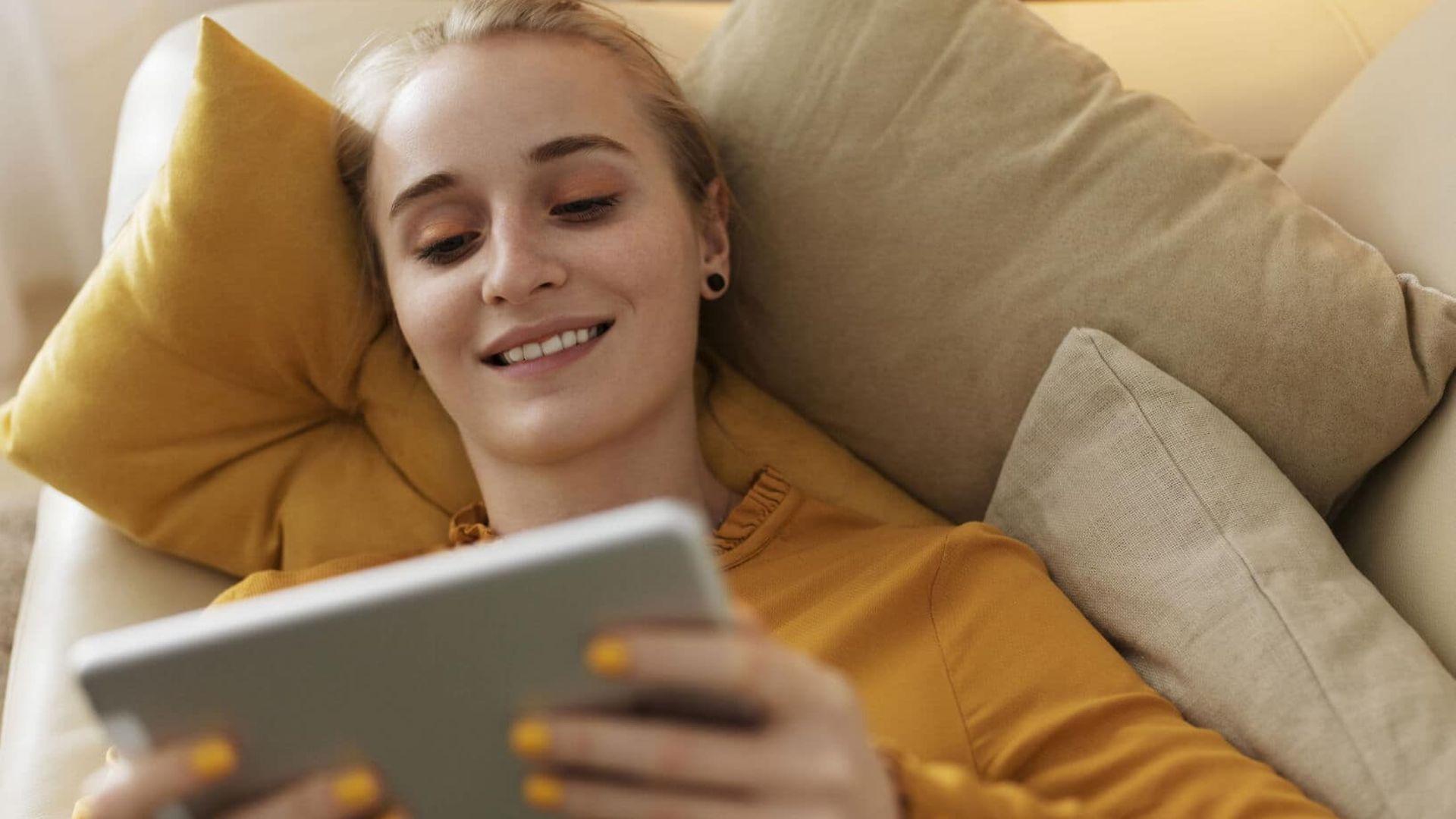 Symbolfoto elektronische Patientenakte: Frau liegt auf Sofa und schaut lächelnd auf Tablet.