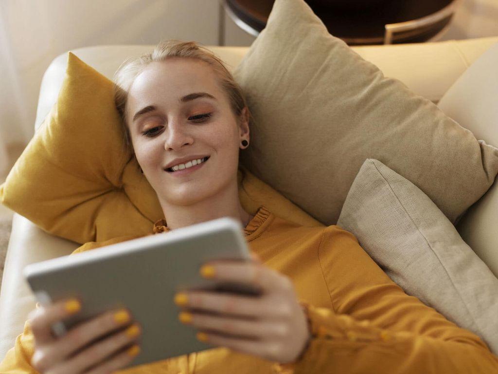 Symbolfoto elektronische Patientenakte: Frau liegt auf Sofa und schaut lächelnd auf Tablet.
