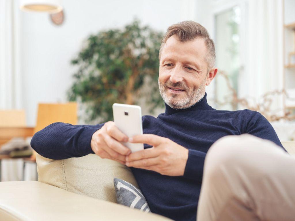 Symbolbild: Mann sitzt auf seiner Couch und lädt mit seinem Handy die E-Rezept App herunter.