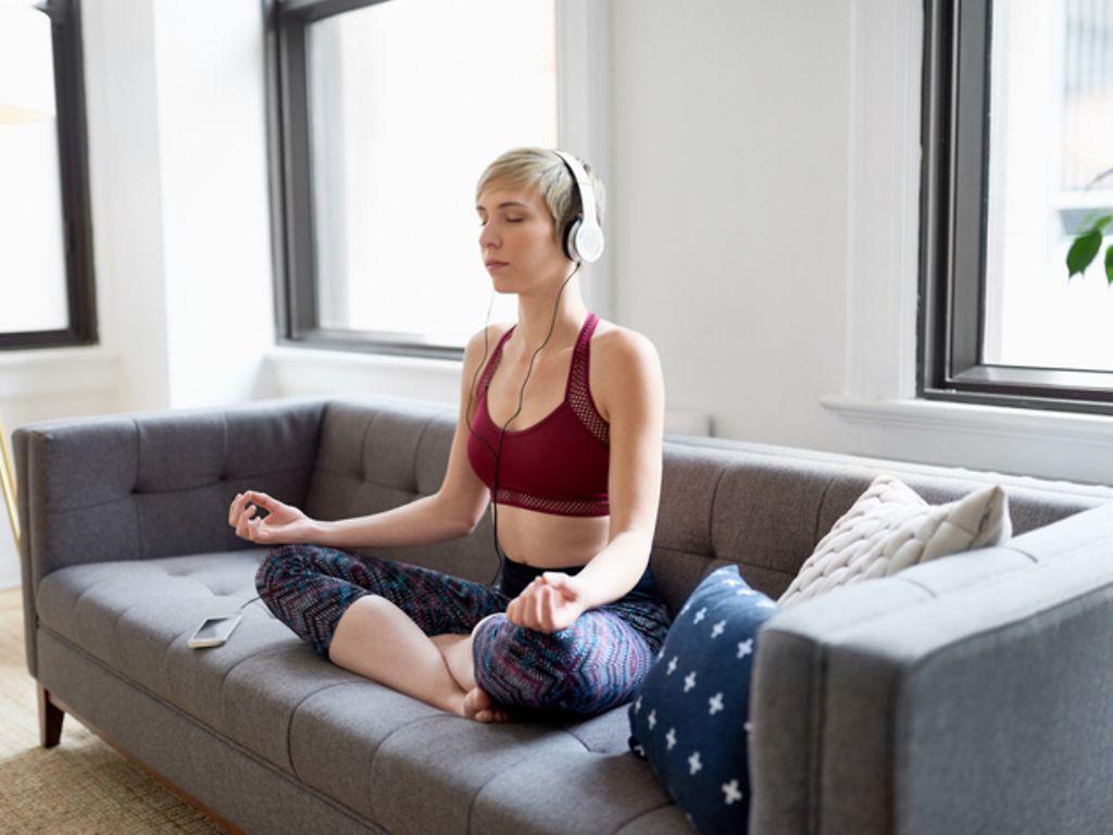 Körper & Seele: Eine junge Frau sitzt auf einem Sofa und meditiert 