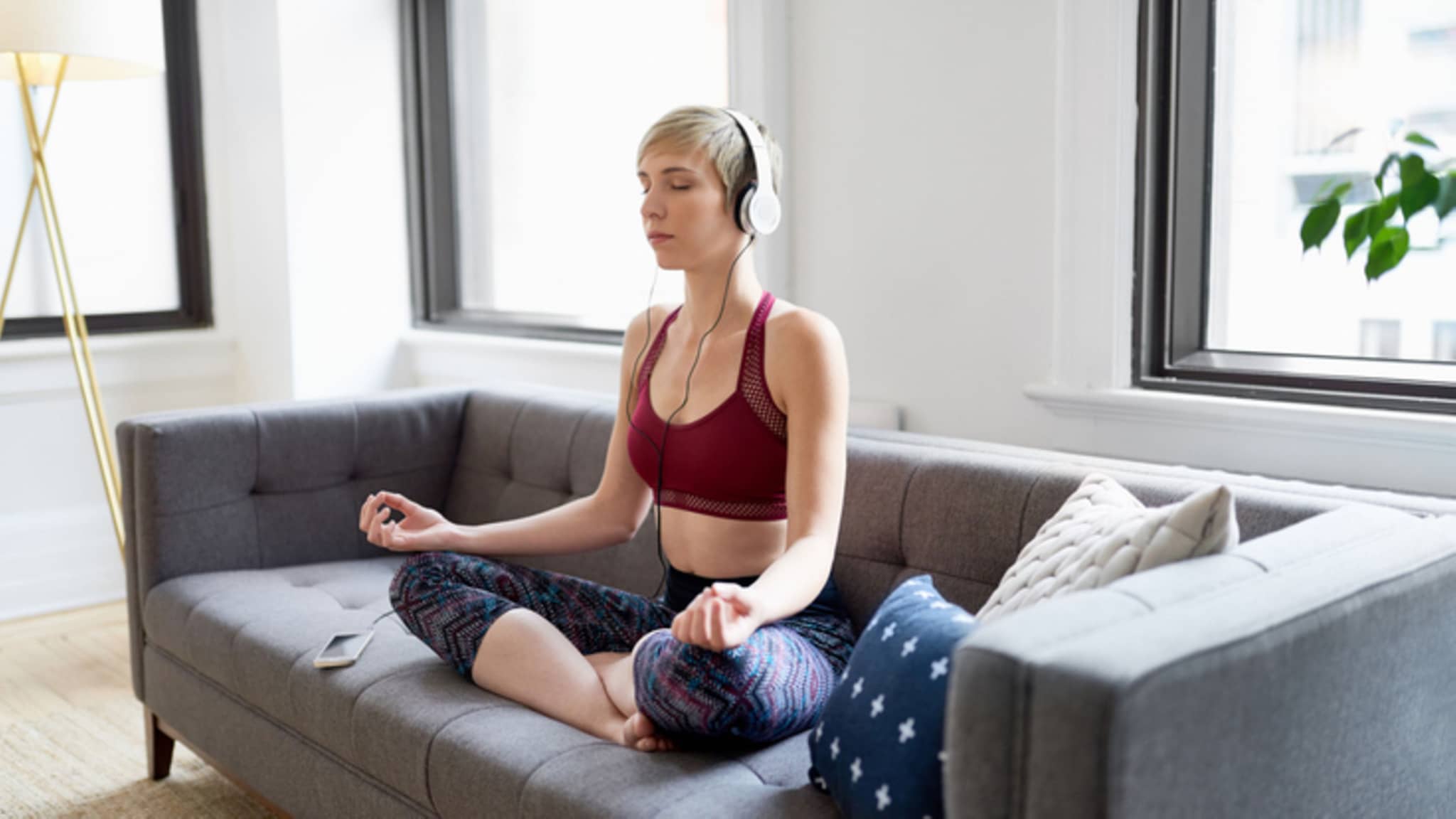 Körper & Seele: Eine junge Frau sitzt auf einem Sofa und meditiert