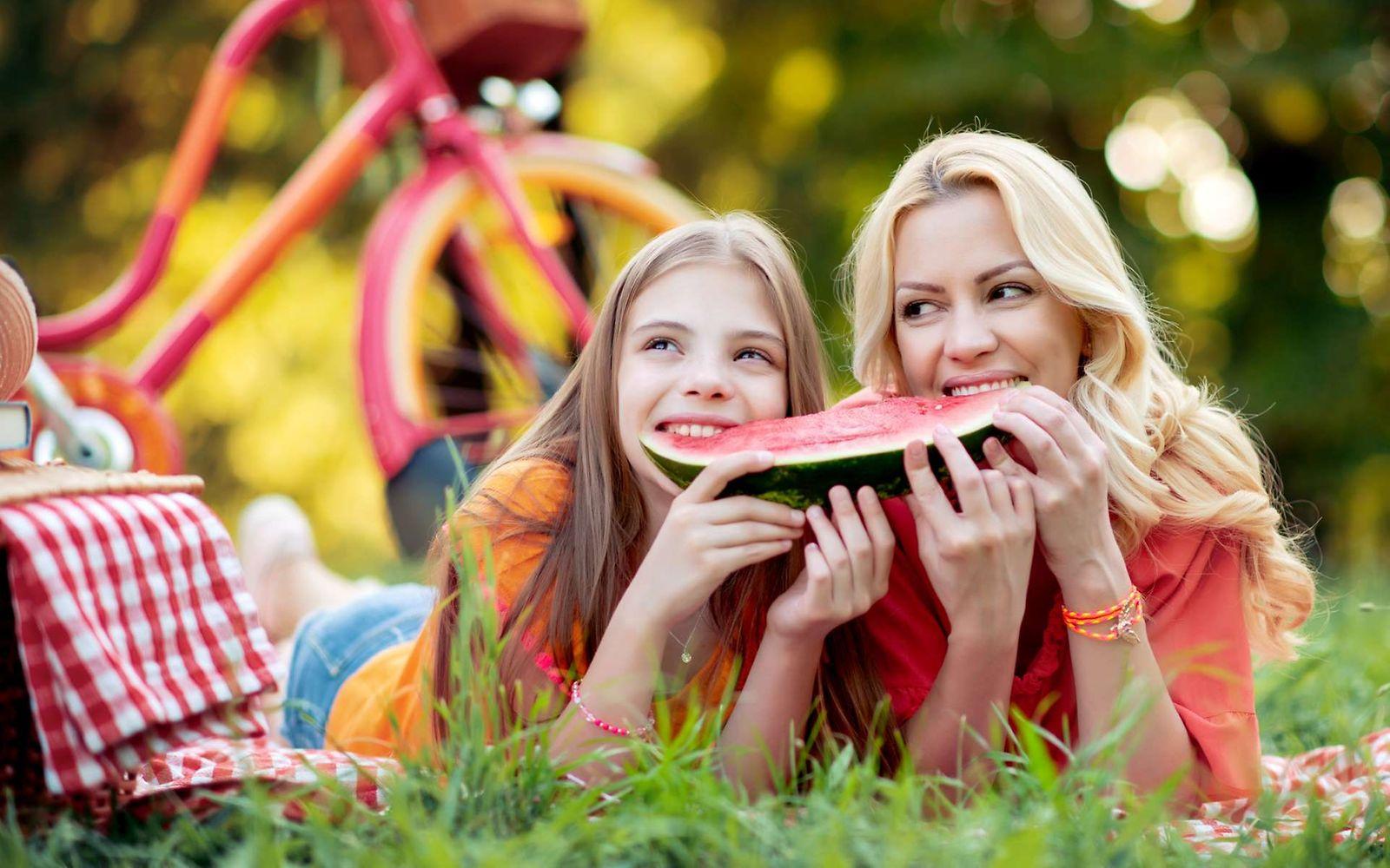 Ernährung bei Hitze: Mutter und Tochter liegen im Sommer im Park und essen eine Wassermelone.