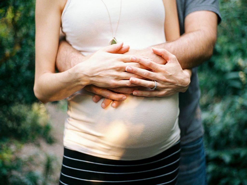 Dammschnitt: Mann umfasst schwangere Frau von hinten und legt seine Hänge auf ihren Bauch