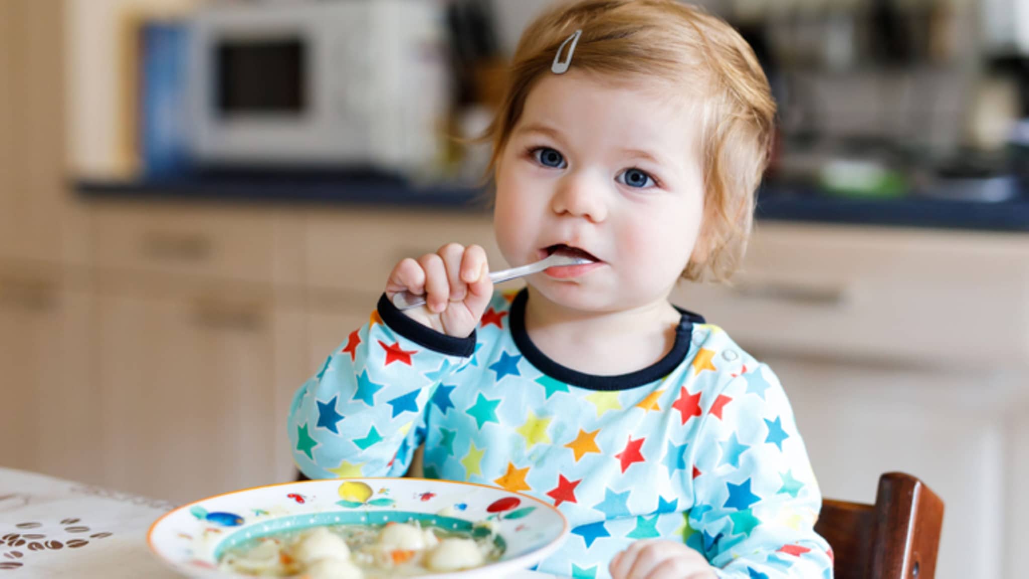 Baby- und Kinderernährung: Kleines Kind sitzt auf dem Hochstuhl mit einem Löffel im Mund.