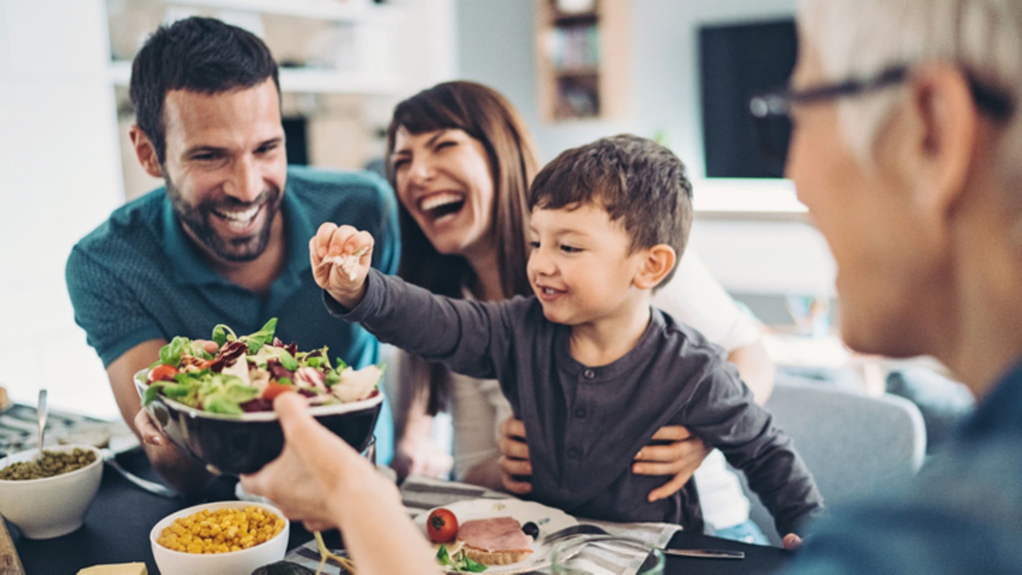 Ernährungstipps: Fröhliche Familie mit Kind sitzt am Tisch und isst Salat.