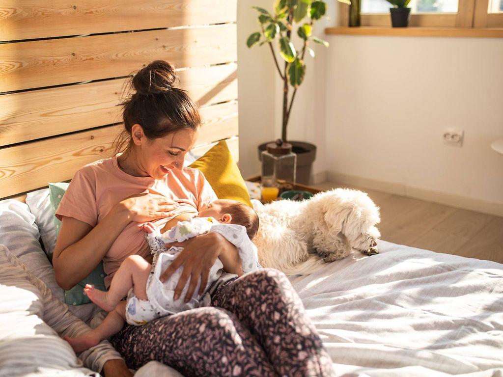 Stillen oder Flasche?: Mutter stillt im Bett ihren Säugling und lächelt entspannt