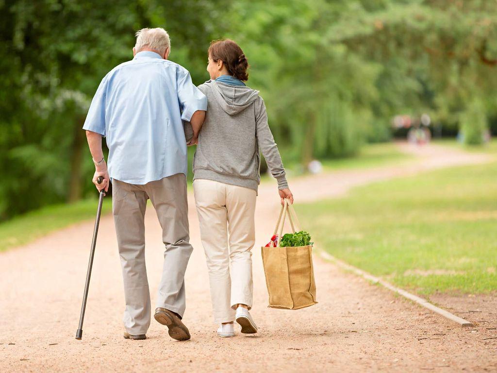 Pflege zu Hause: Junge Frau stützt älteren Mann beim Laufen im Park