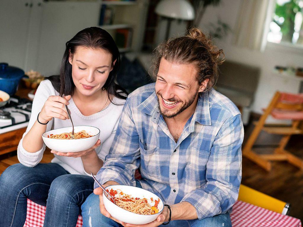 Kohlenhydrate: Glückliches Paar sitzt auf dem Tisch und löffelt Nudeln aus Schälchen