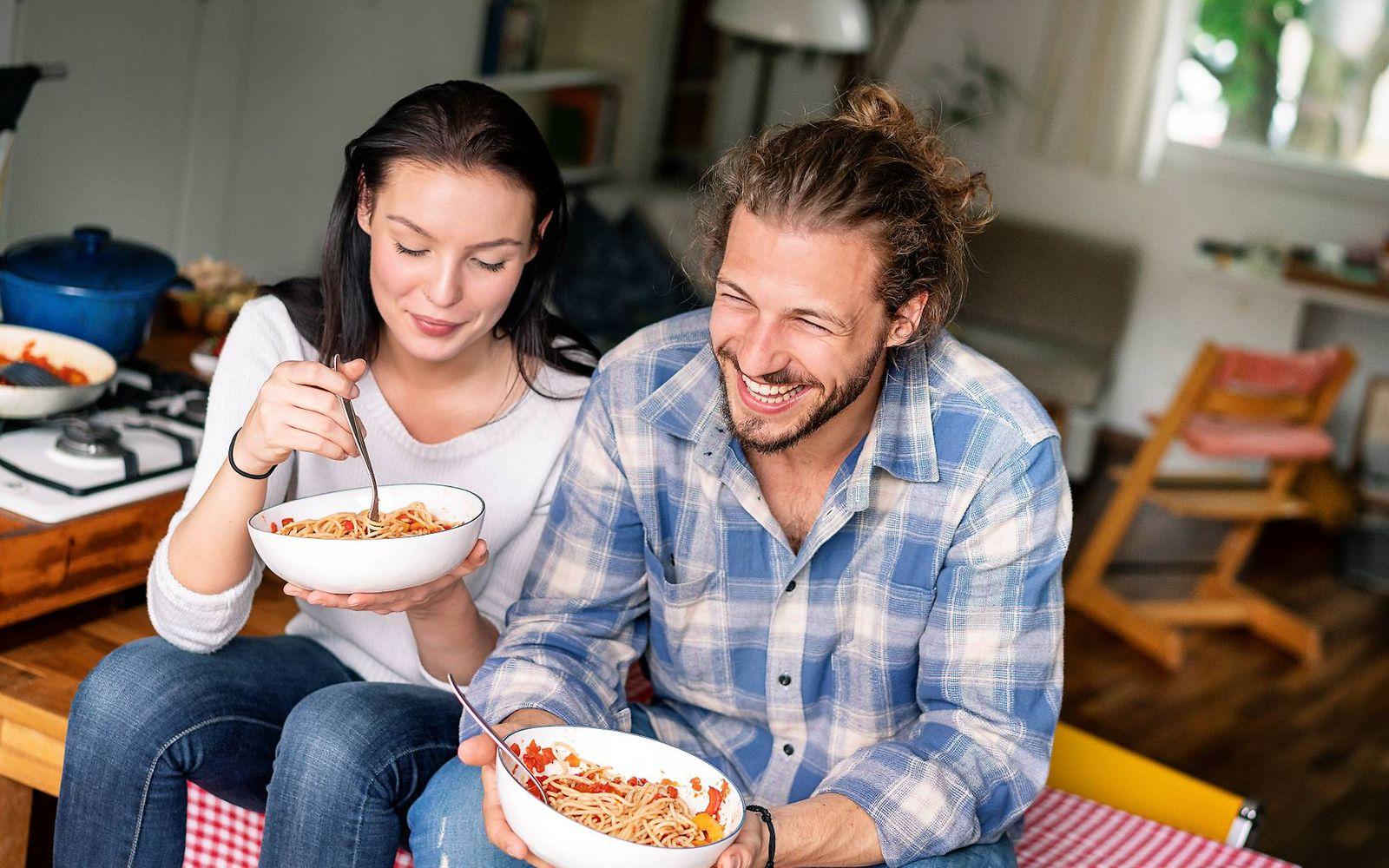 Kohlenhydrate: Glückliches Paar sitzt auf dem Tisch und löffelt Nudeln aus Schälchen