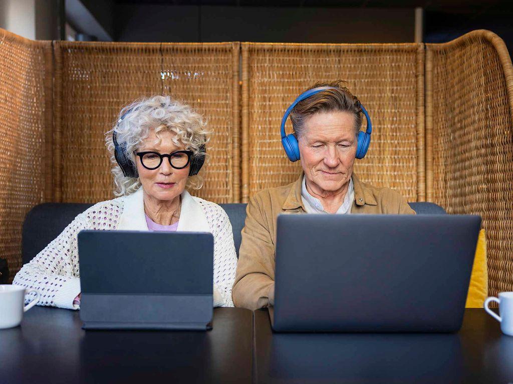 Bild: Ältere Frau und älterer Mann sitzen mit Kopfhörern vor Laptops.