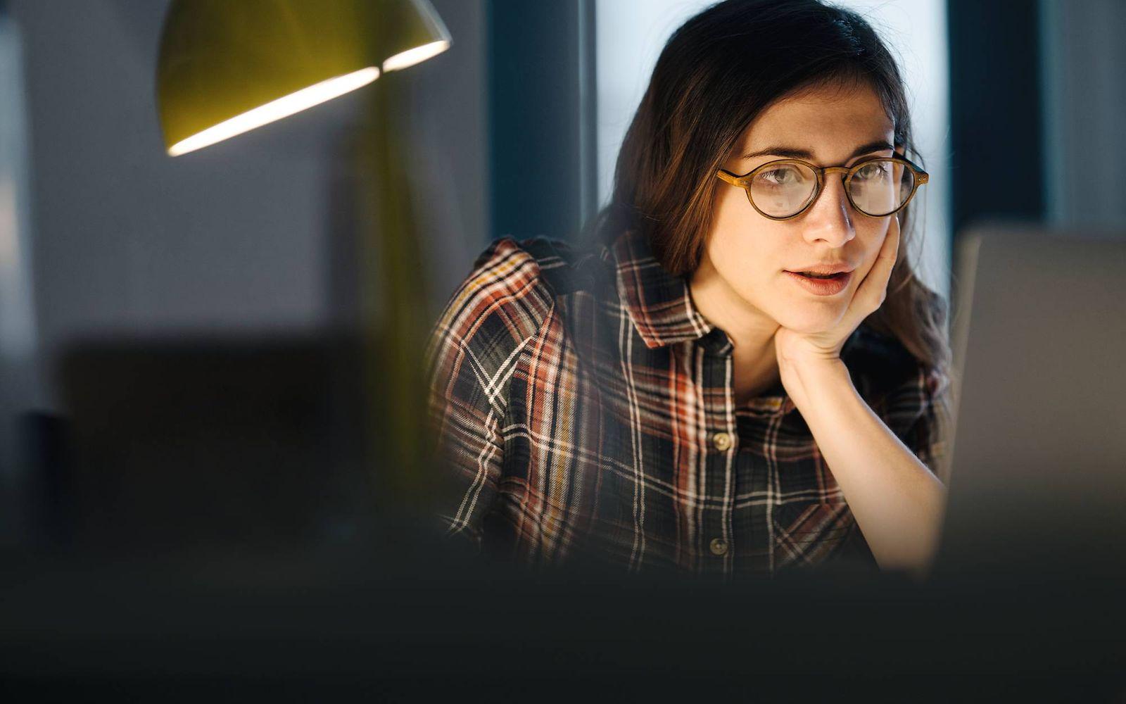 Bild: Junge Frau mit Brille liest etwas auf dem Laptop im Arbeitszimmer.