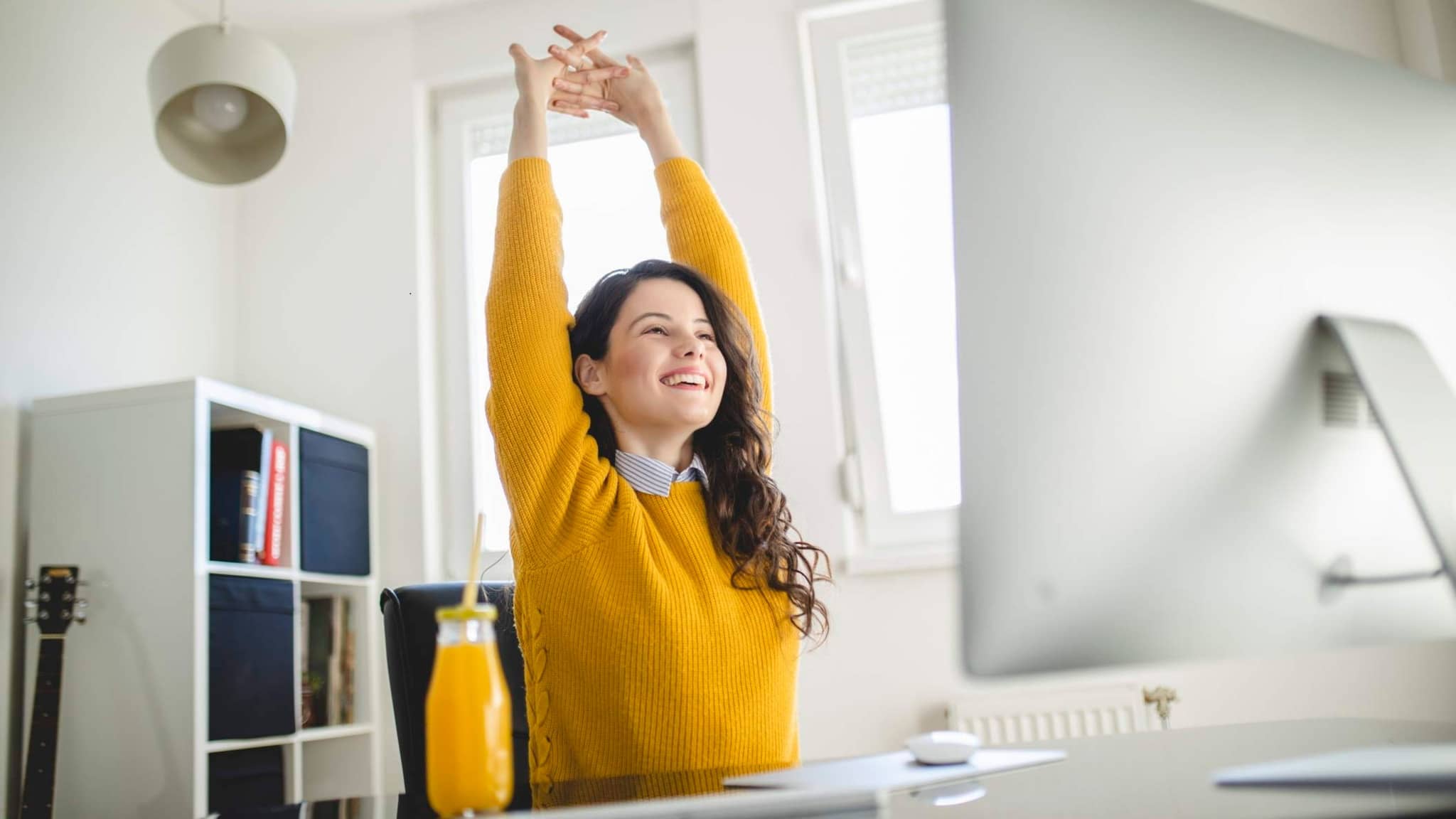 Bild: Frau mit gelben Pullover streckt die Arme nach oben am Schreibtisch.