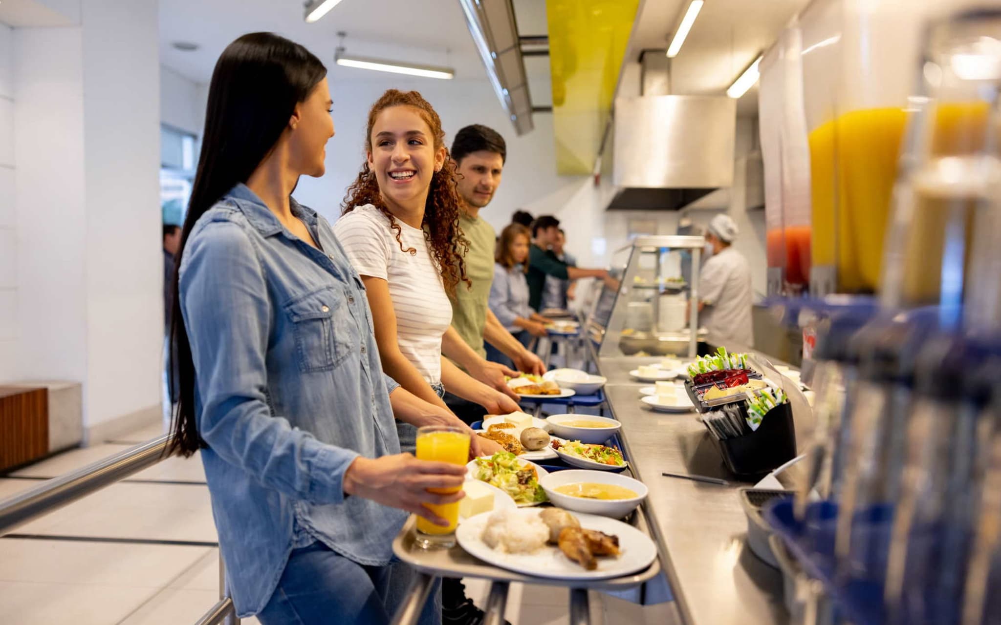 Bild: Menschen stehen in der Kantine mit ihren Tabletts und erhalten Essen.