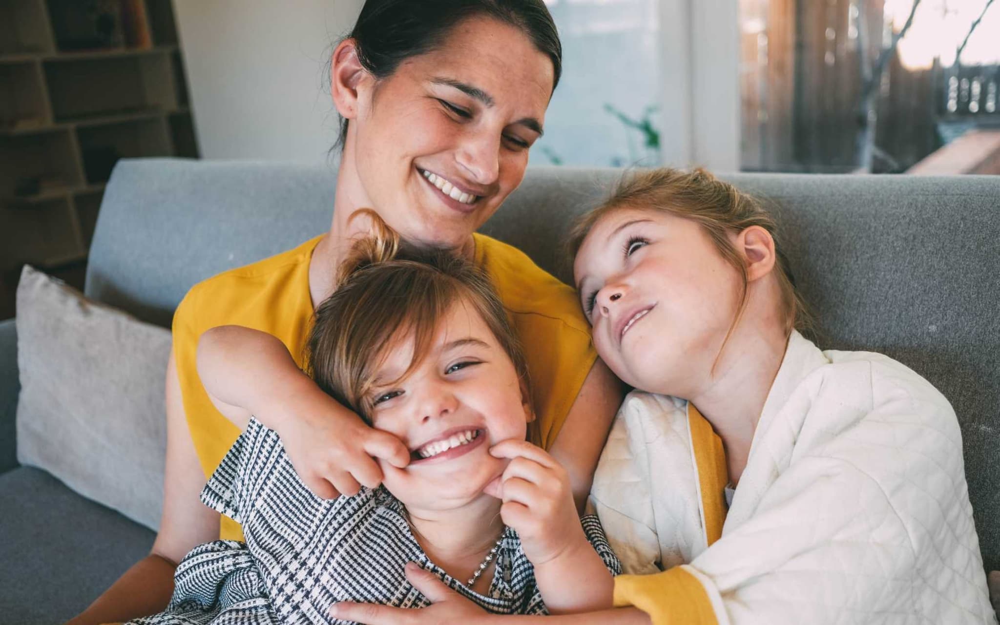 Bild: Mutter mit zwei kleinen Töchtern kuscheln auf dem Sofa und freuen sich über die Vorteile bei der DAK.