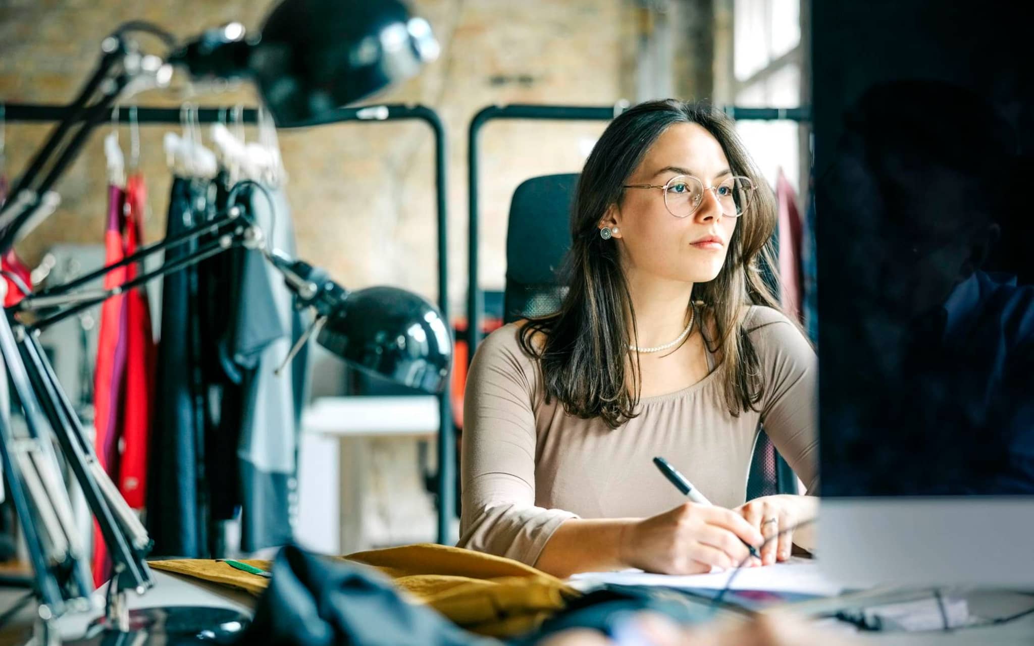 Bild: Frau sitzt an einem Schreibtisch, hinter ihr sind Kleiderstangen mit Kleidung.