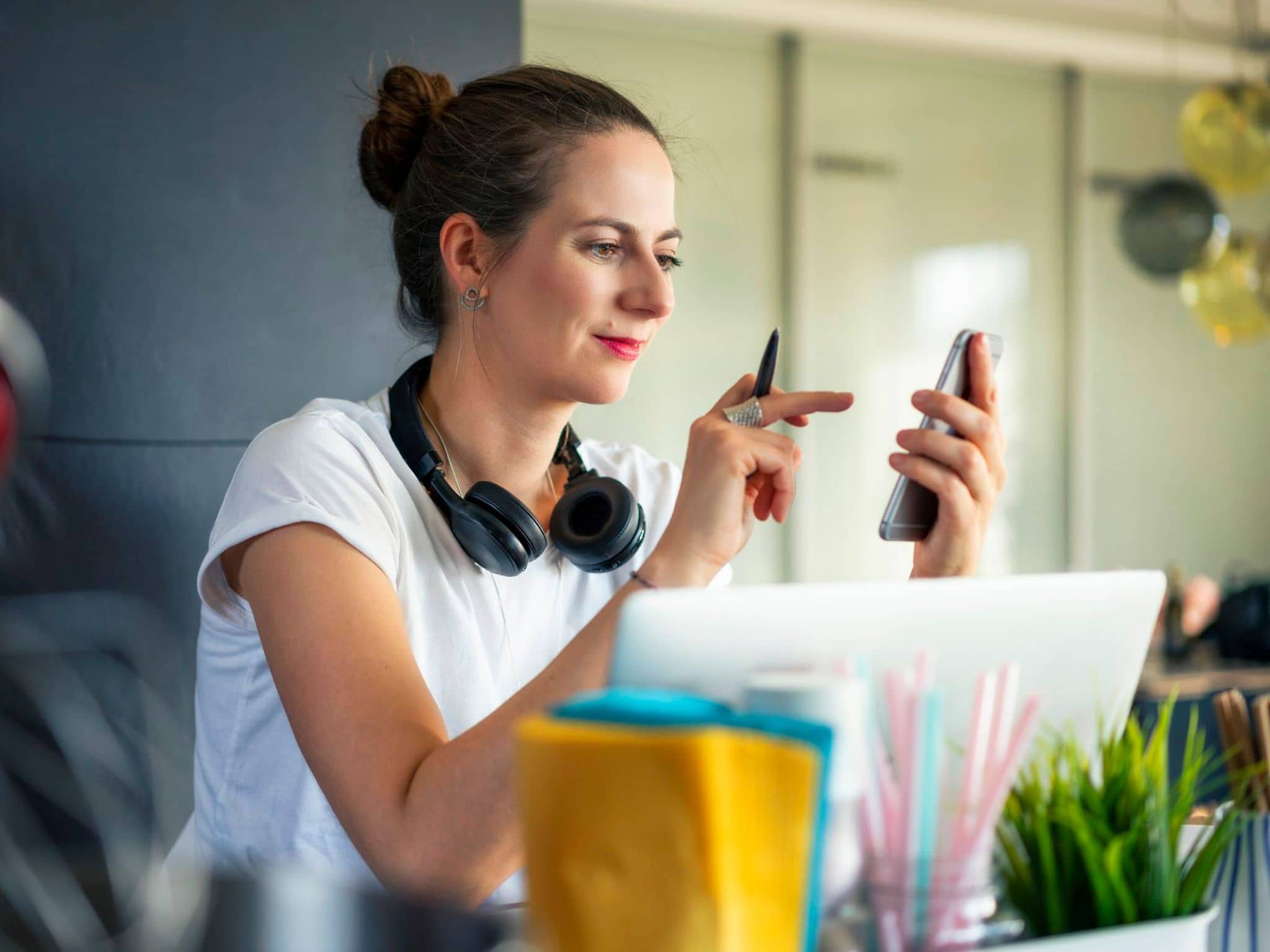 Person mit hochgestecktem Haar und Kopfhörern hält Smartphone und Stift.