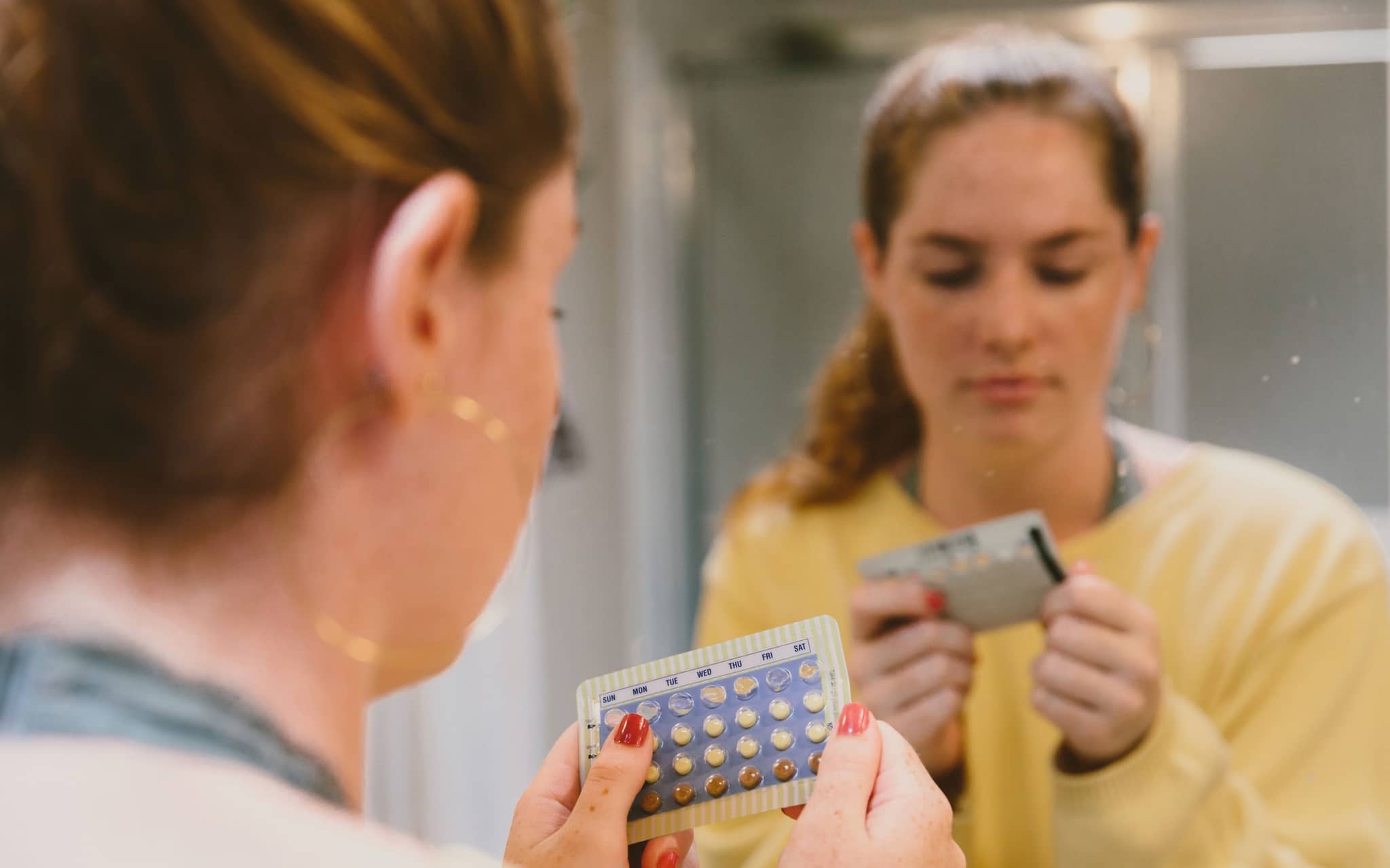 Symbolbild Pillenarten: Eine junge Frau steht vor einem Spiegel im Badezimmer und betrachtet eine Pillenpackung