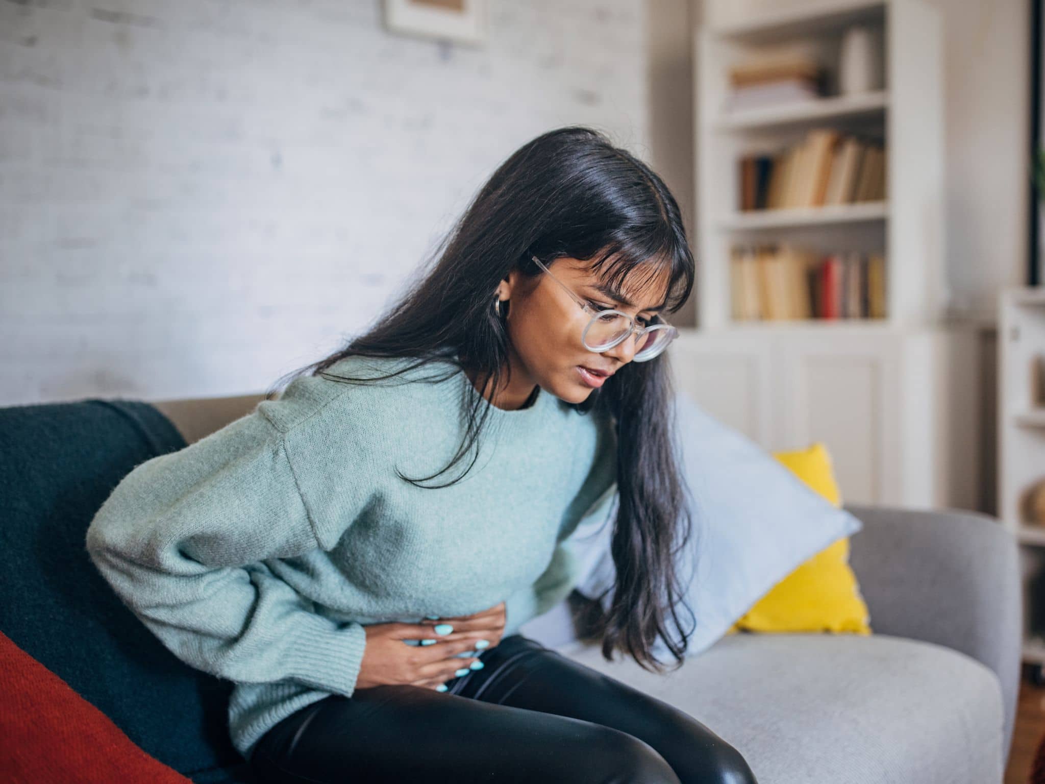 Eine junge Frau sitzt auf einem Sofa und hält sich mit schmerzverzerrtem Gesicht den Bauch.