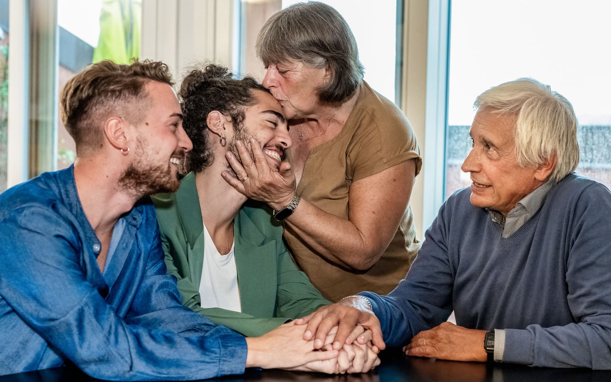 Symbolbild für Outing: Zwei Männer sitzen am Esstisch bei den Eltern, die Mutter küsst einen der Männer liebevoll auf die Stirn