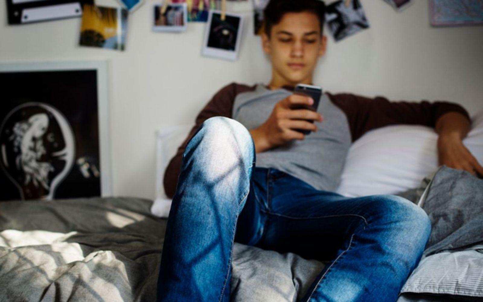 Symbolbild Pubertät bei Jungen: Ein Teenager sitzt auf einem Sofa und guckt auf ein Smartphone