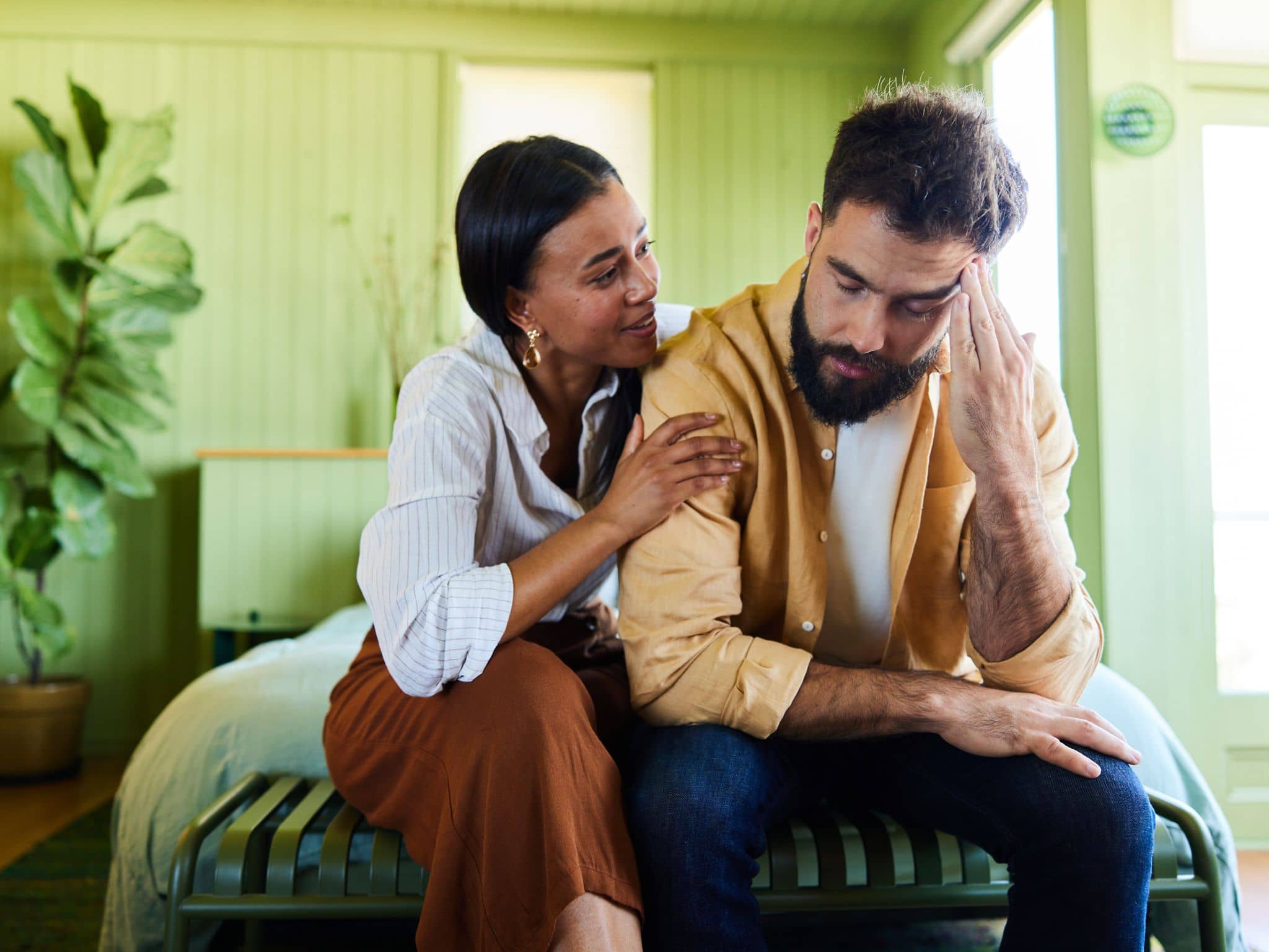 Ein junger Mann mit Vollbart sitzt am Rand eines Bettes, guckt bedrückt und wird von einer Frau getröstet.