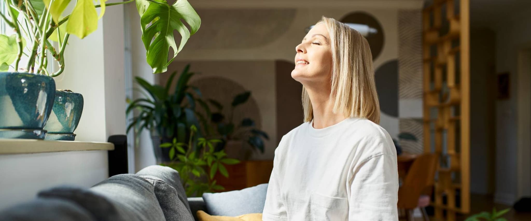 Osteopathie-Zuschuss: Zufriedene Frau sitzt auf dem Sofa und hält das Gesicht in die Sonne.