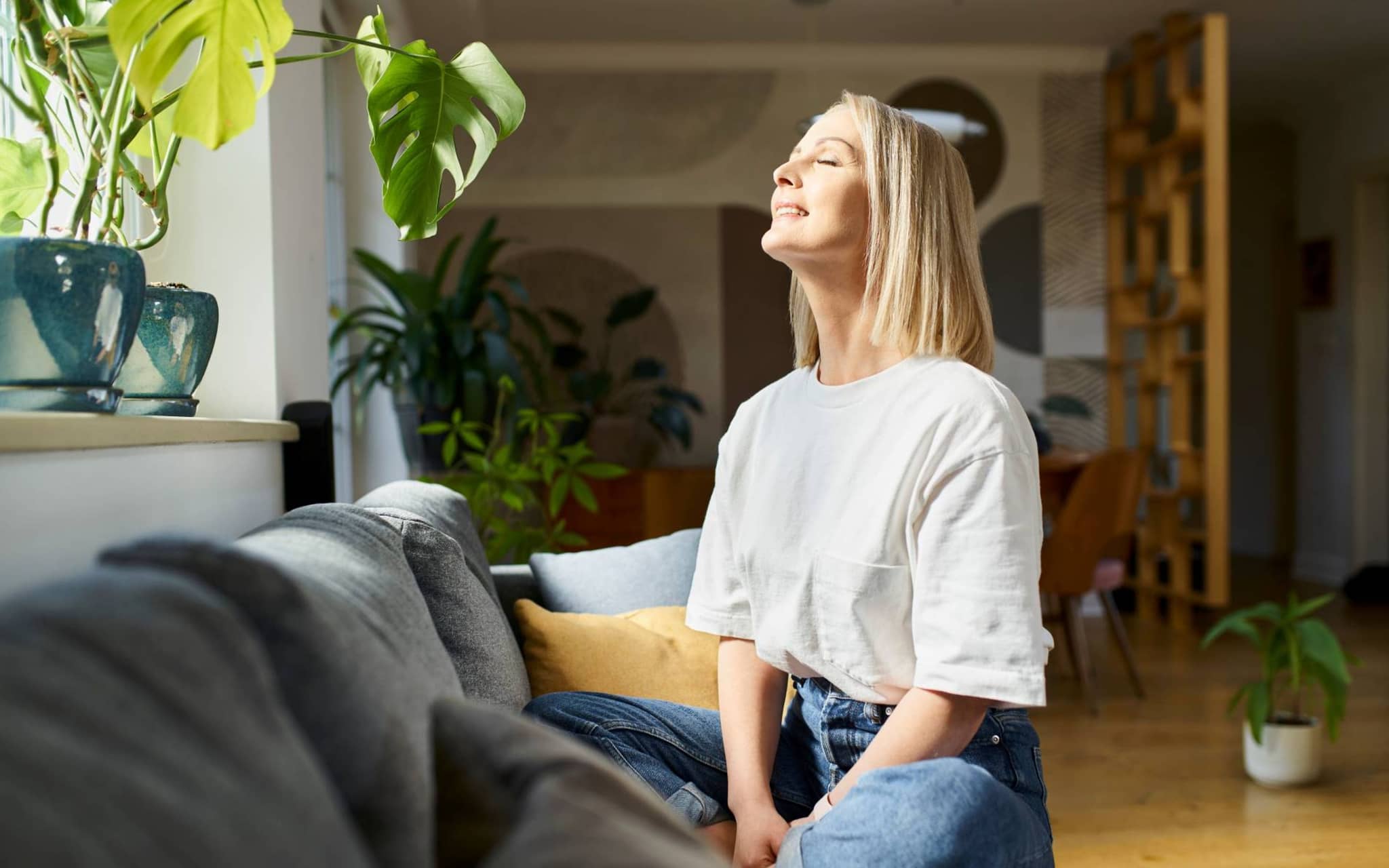 Osteopathie-Zuschuss: Zufriedene Frau sitzt auf dem Sofa und hält das Gesicht in die Sonne.