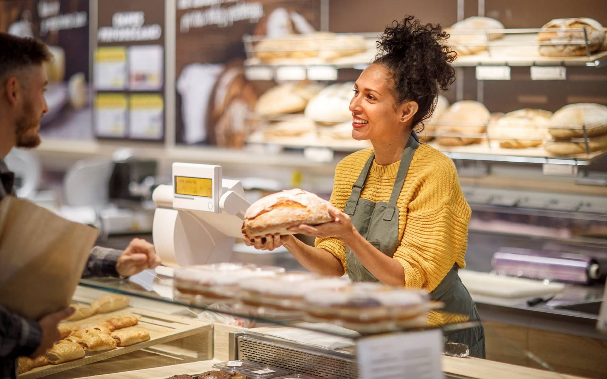 Krankenversicherung im Minijob: Freundliche Bäckereiverkäuferin präsentiert einem Kunden Brotlaib